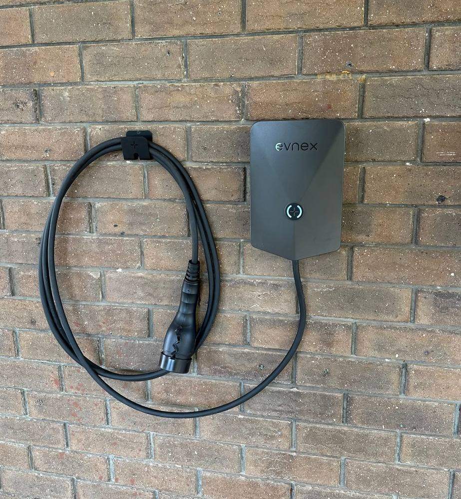 A black Evnex wall-mounted EV charger installed on a home brick wall, with its tethered charging lead coiled neatly on a wall hook beside it