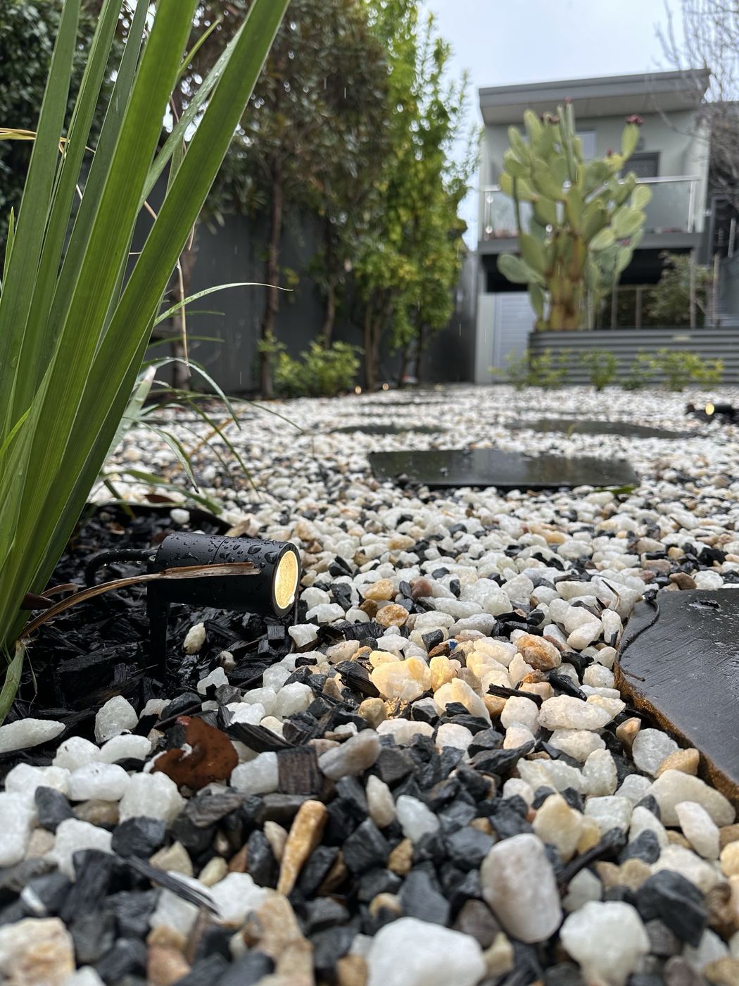 Black garden spotlight set among white pebbles, glowing in the rain